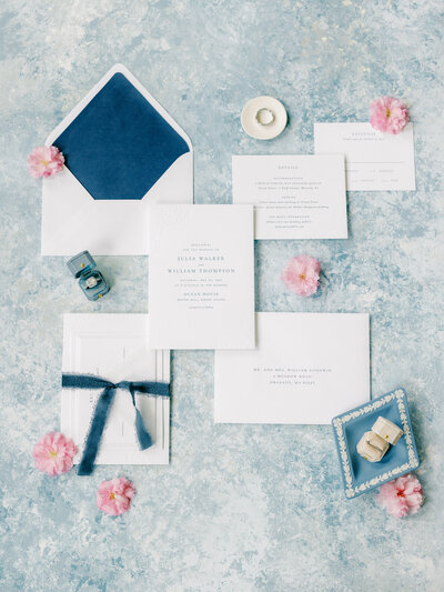 Flay lay of a white and blue themed wedding invitation suite and wedding rings surrounded by pink flowers and rings on a blue and white background