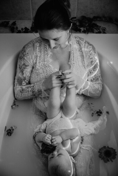 Black and white photo of mom in bathtub holding babys feet and smiling down at baby with flower.