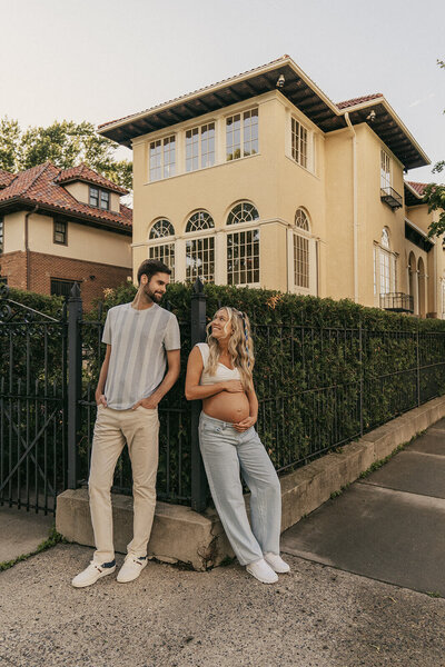 Pregnant woman cradles her stomach while looking up at her husband as they meander down the streets of greater Minneapolis