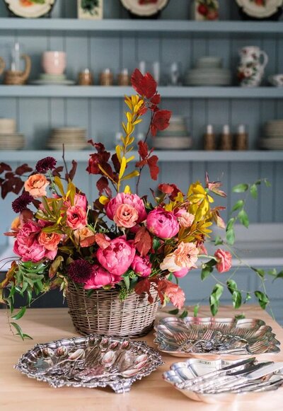 Pink floral arrangement in a basket on a table with a blue bookshelf with dinnerware in the background