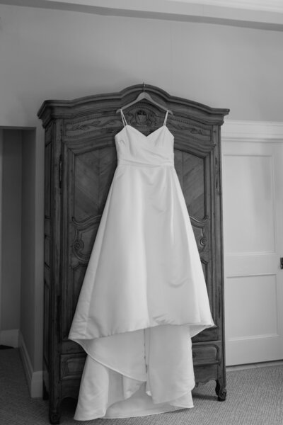 An elegant wedding dress hangs on a dark, ornate armoire, waiting to be worn, the dress and furniture together creating a scene of anticipatory beauty.