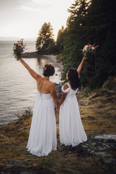 lesbian newlywed couple celebrating by the water at sunset with their bouquets in the air