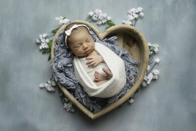 A beautiful newborn girl, wrapped in soft fabric, peacefully laying in a heart-shaped arrangement of flowers, creating a serene and delicate moment, captured by Chunky Monkey Photography.