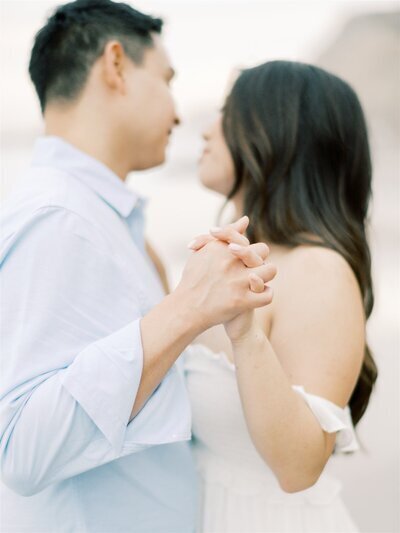 Kohnur Bride with soft glam makeup and effortless loose waves embraces her groom in a sunlit hillside field, capturing a romantic and natural engagement moment with timeless wedding elegance