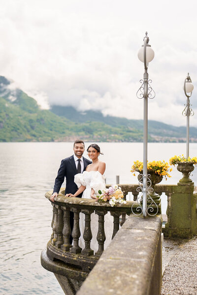 man and woman standing at stone wall next to water with mountains in the background
