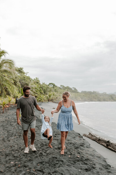 A family of three all holding hands walk along the black beach in Puerto Viejo Costa Rica having a great photoshoot with sierra bertini photography