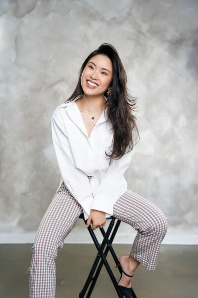 Stephy Chen sitting on a stool in a white blazer and checkered pants, smiling confidently during a modern branding shoot.