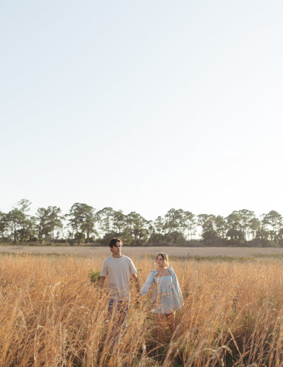 two people holding hands walking towards the camera through tall golden grass