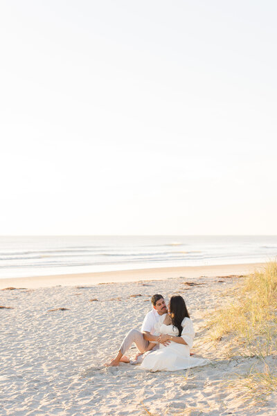 Pregnant couple sit together near the dunes with the ocean in the background during their session with an Orlando maternity photographer