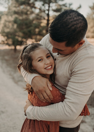 A dad looks lovingly at his daughter as he wraps his arms around her, and she gives iContact to the camera while she laughs.