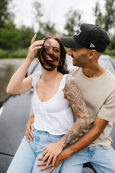 A man and woman sit on a car hood, smiling and laughing. The woman, showcasing tattoos, wears a white top and jeans. The man sports a cap and beige shirt. Trees frame the joyful moment, captured perfectly by Toronto engagement photographer, Jennifer van Son.
