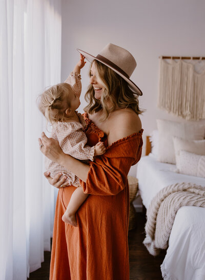 Mother in a pretty rust boho dress holding her toddler who reaches for her hat.