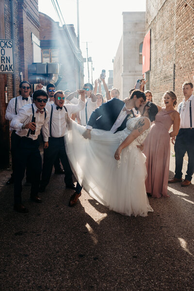 A groom dipping his bride as their bridal party yell and celebrate next to them.