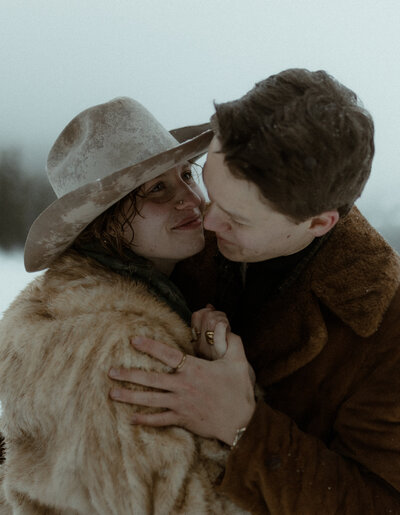 couple embracing closely in a snowy winter landscape, dressed in warm vintage-inspired coats. The woman, wearing a wide-brimmed hat dusted with snow and a fur coat, gazes up at her partner with a soft smile. The man, wrapped in a brown shearling jacket, leans in tenderly with a loving expression.
