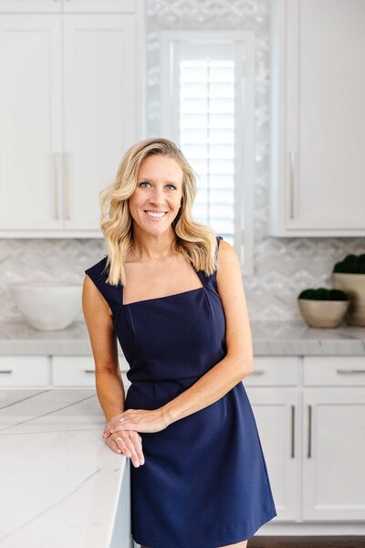 A Frisco interior designer in an elegant blue dress, standing next to a beautifully renovated white kitchen.