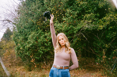 A Toronto family photographer with long blonde hair in a beige top holds a camera aloft while standing in front of dense green foliage.