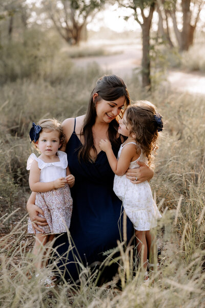 mom in a elegant navy blue dress sitting in a grass field with a pathway lined with trees behind her. She is smiling down at her older daughter as she hugs both girls .