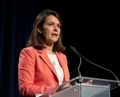 Leslie Hansen delivering a keynote speech at a podium in a coral blazer.