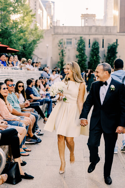 A bride and groom walking hand in hand along the Chicago Riverwalk with city skyscrapers in the background.