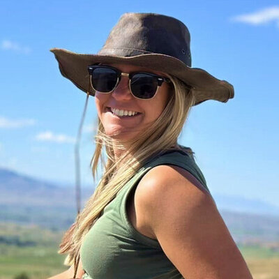 Woman with blonde hair smiling in the plains, wearing a wide-brim hat, sunglasses and tanktop.
