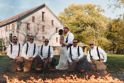 Bride and groom kissing in front of groomsmen kneeling with fire at the bottom. A barn is in the background.