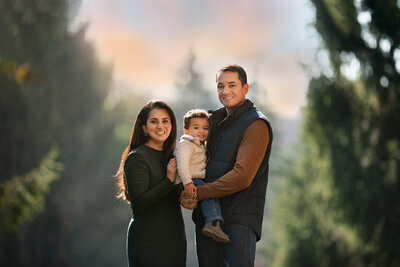 Family of 3 standing with pine trees in the background photo taken by Southeast Michigan Photographer Kat Figlak