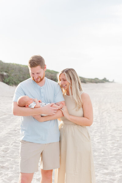 New parents hold their newborn baby boy on the beach and smile at him during golden hour newborn session