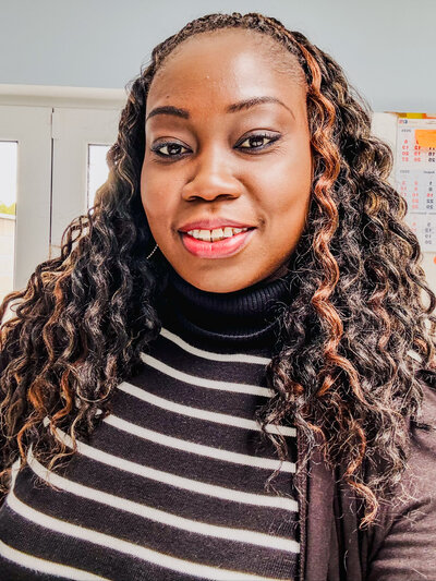 A professional portrait of a confident Black woman with shoulder-length twist braids and a warm smile. She is wearing a black turtleneck sweater and gray high-waisted pants. The background features a modern office environment with large windows showing an urban view.