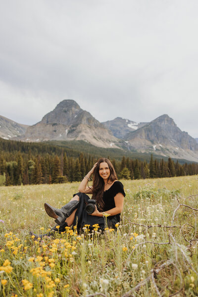 Aurelia, Montana wedding photographer, sitting in a flower field with her camera, smiling in front of the mountains.