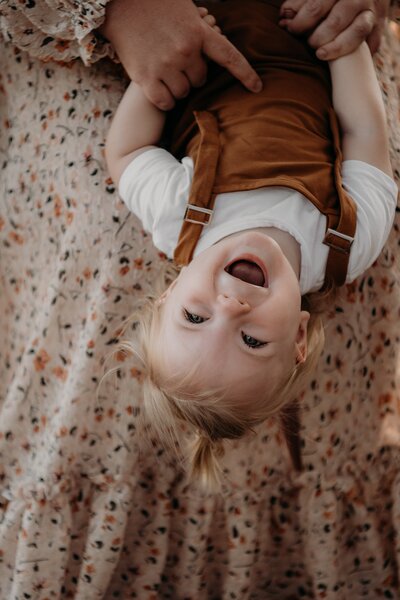 A toddler with blonde hair and pigtails, wearing a white shirt and brown overalls, is smiling while being held upside down by an adult—captured perfectly by NSW family photographer.