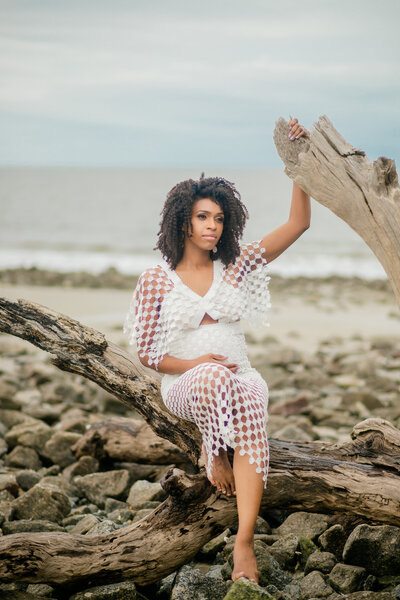 A beautiful Black woman wearing a white knit dress sits on driftwood at Driftwood Beach, showcasing her pregnancy. Makeup by TamGee Artistry enhances her glowing beauty.