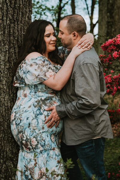 Pregnant wife wearing a blue floral dress smiling at husband wearing gray while he is holding her belly in flower garden in Annapolis Maryland.