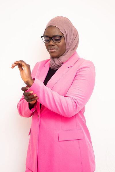 Photo of Isatou Bittaye Jobe, a grant writing expert, dressed in a pink blazer, standing against a white backdrop. She is wearing a brown head wrap and black glasses. 