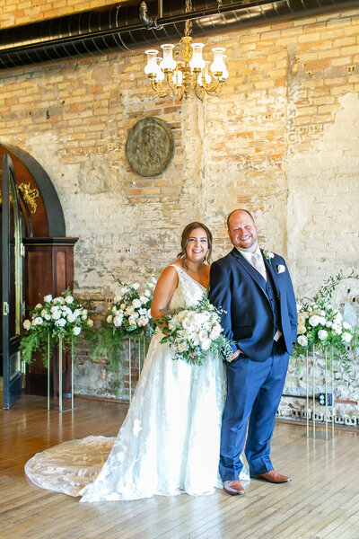 A white couple standing in front of a cream brick wall wearing wedding attire. They are surrounded by florals
