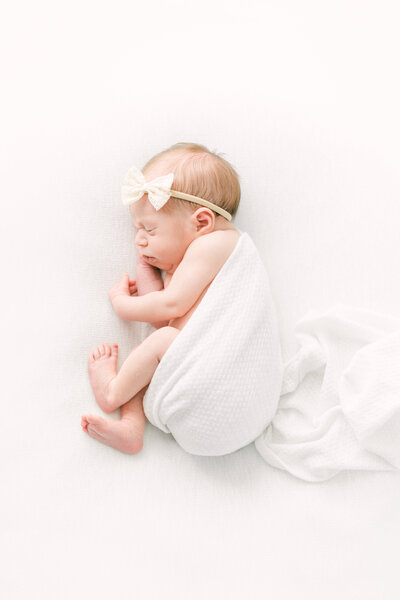 A newborn baby in a white onssie sleeps during photos. 