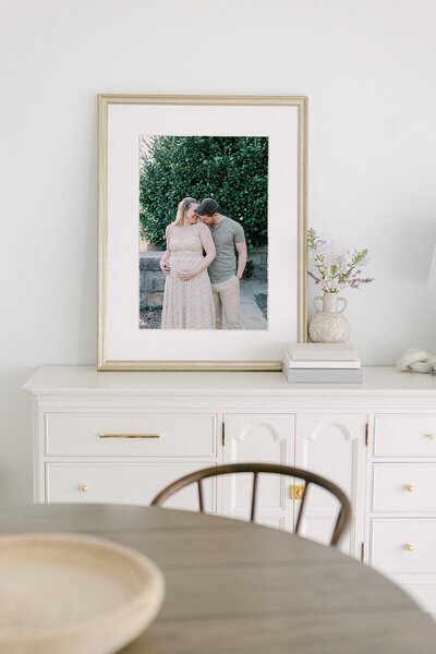 A framed print resting on a white console table with a table and chair in the foreground. The framed print is of a man kissing a woman's shoulder as the woman holds her pregnancy bump by Katie Stansfield Photography, a family photographer in Richmond.