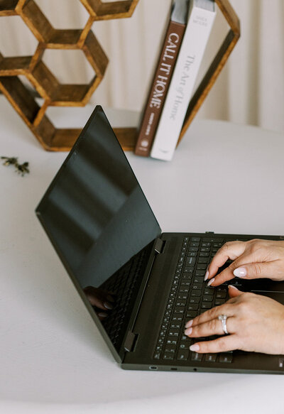 Hands typing on a black laptop on a white table. In the background, there are three stacked books and a hexagonal bookshelf.