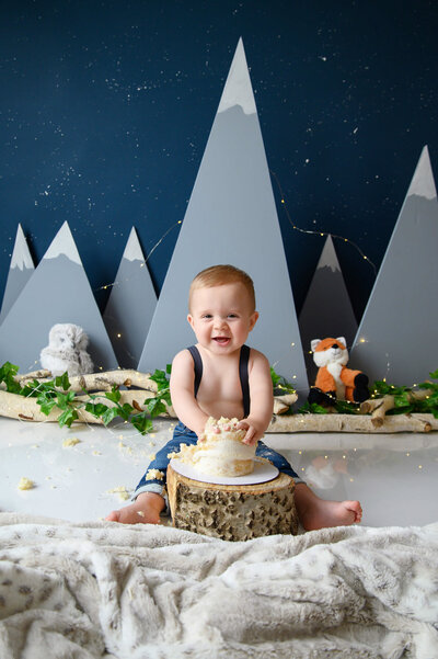 little boy wearing blue jeans with blue suspenders with mountains in the background for his Burlington cake smash photography session.