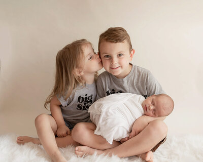 Brother and Sister holding newborn baby brother wrapped in a white swaddle