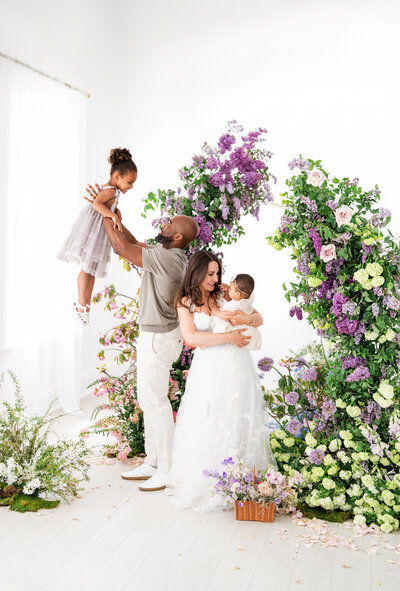 A family of four poses playfully under a floral arch; the father lifts his daughter in a lavender dress, while the mother cradles a baby in cream, surrounded by blooming lilacs, roses, and hydrangeas.