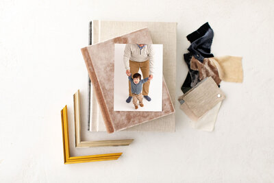 Image of one-year old boy walking while holding hands with his dad behind him. Dad and son are wearing light neutrals with some chambray mixed in. Image is sitting on top of some beautiful heirloom albums and surrounded by corner frames and album cover samples. Milestone baby photographer