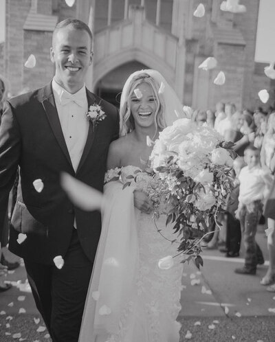 Bride and groom smiling as they walk through falling petals outside a church