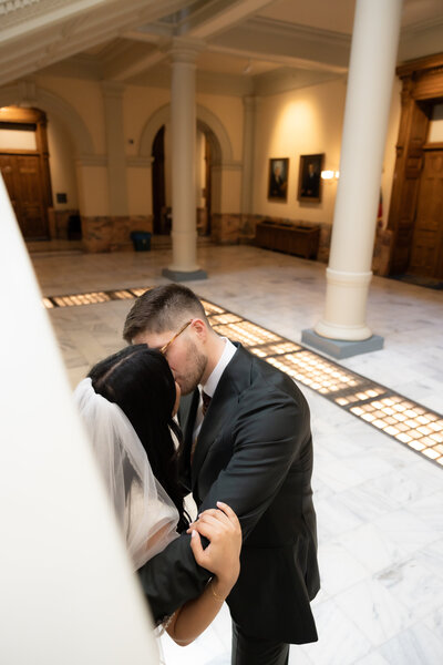 Bride and groom sharing an intimate kiss inside a marble courthouse, framed by white columns and soft indoor lighting.
