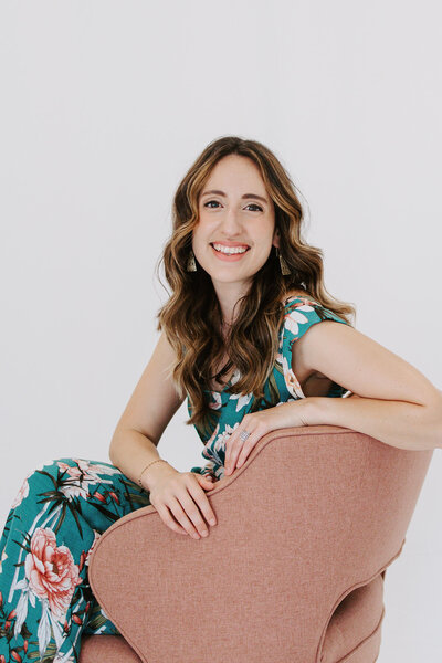 Woman with brown curly hair wearing floral dress and sitting on pink chair