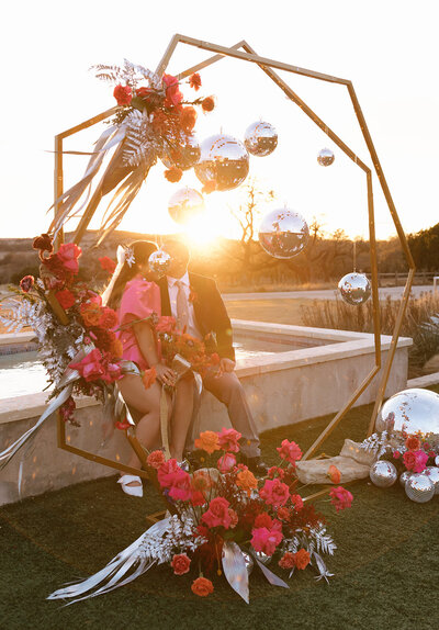 Man and woman sitting under a display arch decorated with disco balls, hot pink flowers, and feathers