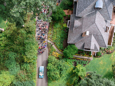 Aerial view of a winding path in a lush garden with a group of people walking in colorful attire. A blue vintage car drives on the path, surrounded by green vegetation and a large house on the right.