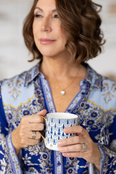 Woman with short wavy brown hair wearing a blue patterned blouse, holding a white and blue mug with both hands and looking off to the side.