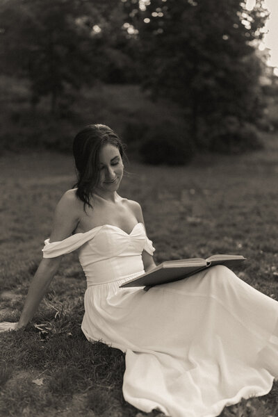 Black and white image of a woman in a white dress sitting on the grass and reading a book