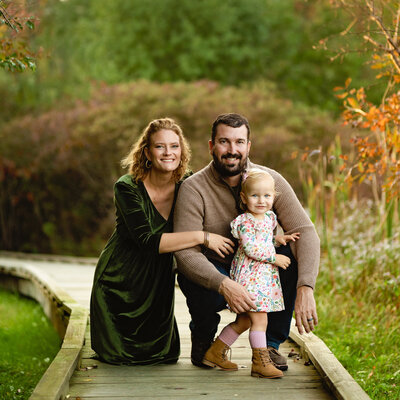 A mom, dad and toddler girl are crouched and posing on a walking trail in the Hudson Valley NY