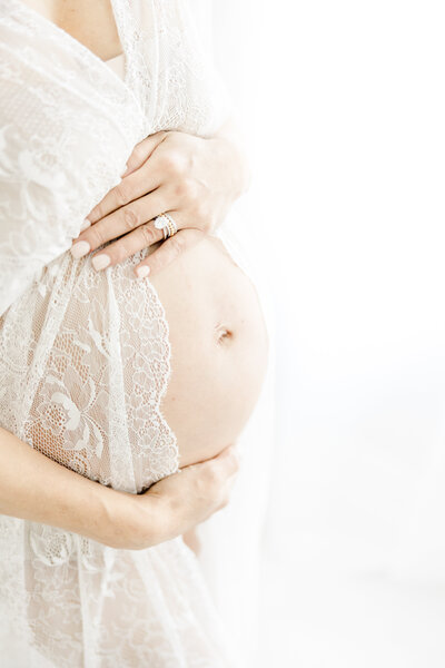 Woman embraces her pregnant belly in a white lace robe during in-studio maternity portrait session